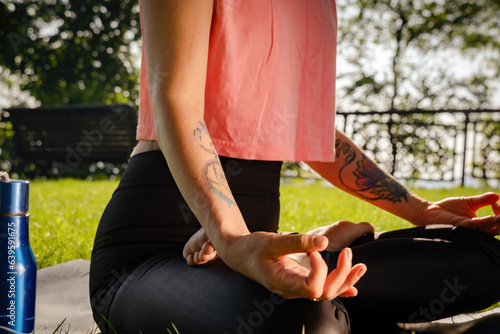 Woman body with tattoo in lotus position against city park. Relaxing meditation on green grass. Mental health care closeup