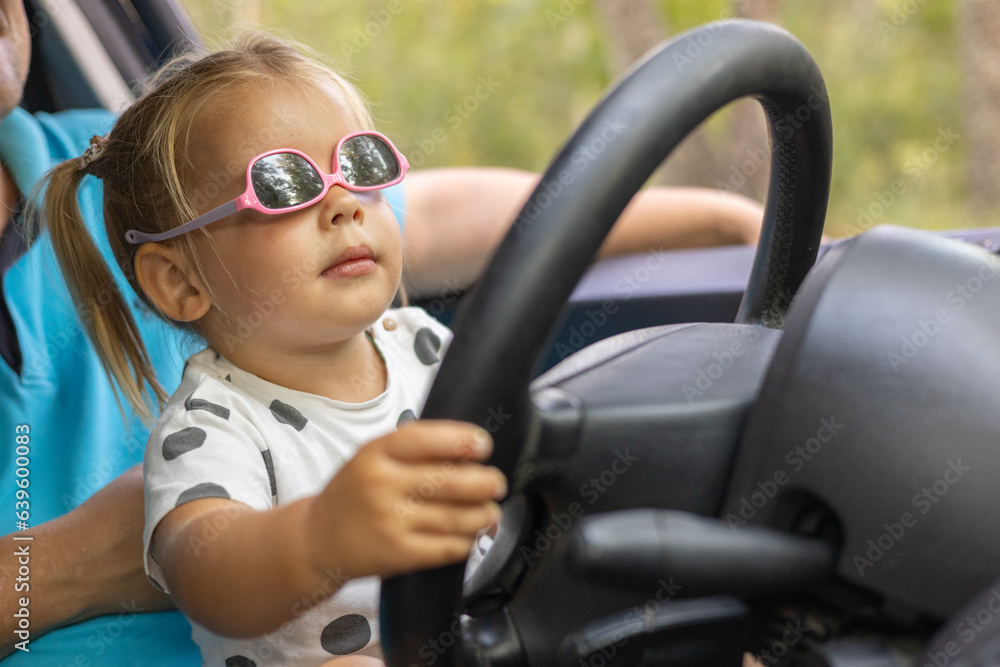 A cute little girl driving a car on her father's lap. Child girl in sunglasses with a serious expression learns to steer a car. A father teaches his daughter how to drive a car. A cute little girl driving a car on her father's lap. Child girl in sunglasses with a serious expression learns to steer a car. A father teaches his daughter how to drive a car.