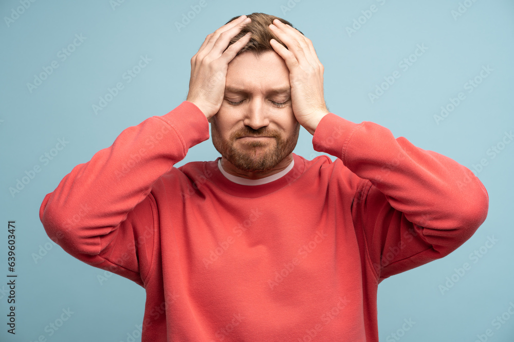 Irritated man with closed eyes holds head feeling discomfort due to headache on blue wall background. Concept of illness and suffering from unpleasant sensations. Sad bearded male regrets a mistake Irritated man with closed eyes holds head feeling discomfort due to headache on blue wall background. Concept of illness and suffering from unpleasant sensations. Sad bearded male regrets a mistake