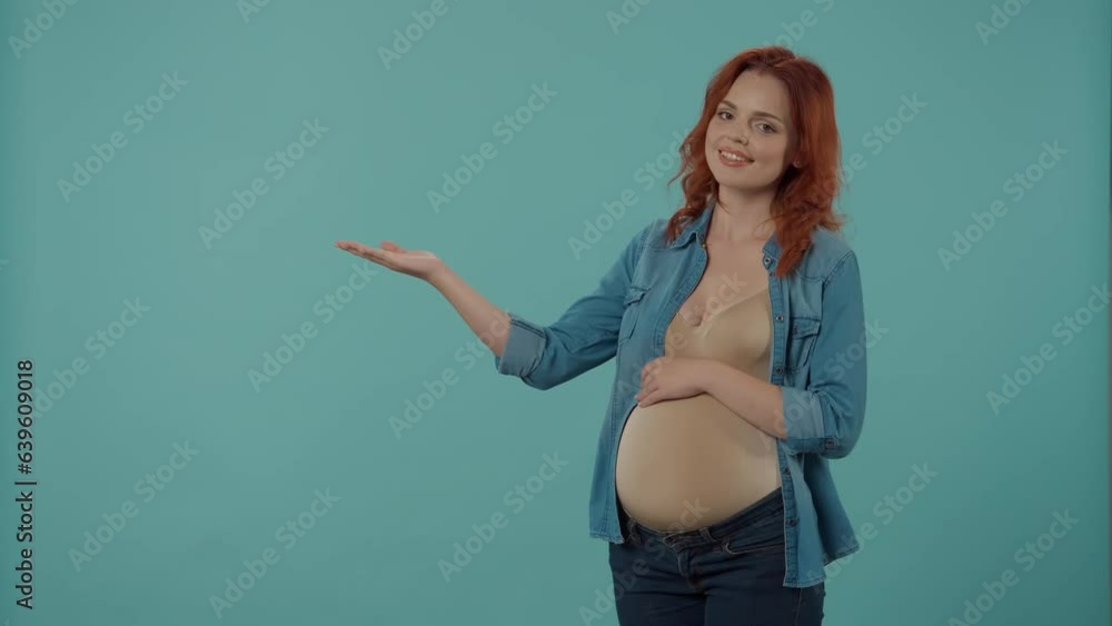 A redhaired woman, expecting a baby, holds a pregnant belly and points with her palm to an empty space for advertising. A pregnant woman nods her head approvingly in the studio on a blue background. A redhaired woman, expecting a baby, holds a pregnant belly and points with her palm to an empty space for advertising. A pregnant woman nods her head approvingly in the studio on a blue background.
