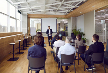 Professional business coach standing by a white board in front of many people in a classroom or office conference room, giving a lecture, and sharing his expertise