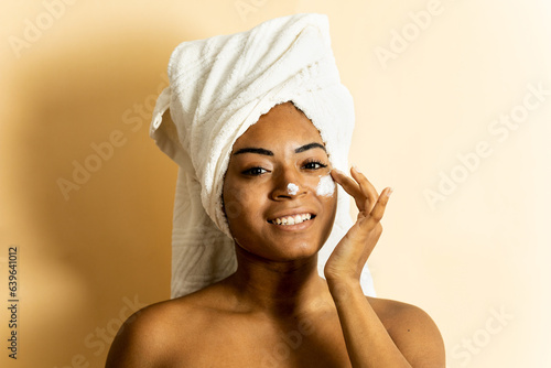 A cute African woman is posing against a brown background while looking at the camera happily. The woman is applying cream to her face while wearing a towel on her head. Applying moisturizer.