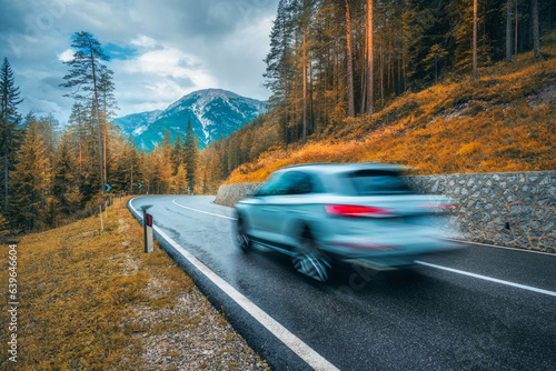 Blurred car in motion on the road in orange forest in rain in autumn. Dolomites, Italy. Perfect mountain road in overcast day in fall. Roadway, pine trees in alps. Transportation. Highway in woods