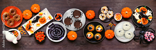 Spooky Halloween treat table scene over a dark wood banner background with copy space. Overhead view. Collection of cookies, cakes, donuts and candies.