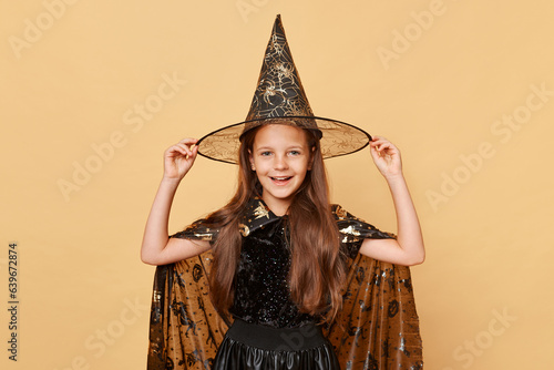 Beautiful little girl wearing witch costume and carnival cone hat isolated over beige background kid sorcerer lookingat camera with happy face at halloween celebration. photo
