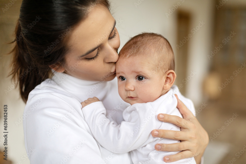 Caring young mom holding and hugging her adorable baby, enjoying motherhood and child care, standing in bedroom at home. Mother's day concept Caring young mom holding and hugging her adorable baby, enjoying motherhood and child care, standing in bedroom at home. Mother's day concept