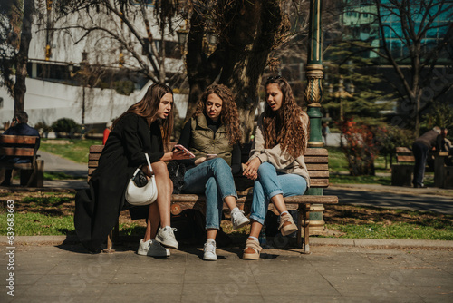 Brunette girl showing something on the phone to her friends. They are sitting on a bench. Front view shot