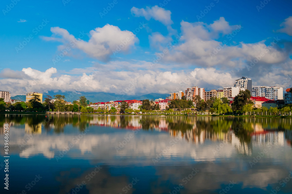 View of the blue sky with white clouds over modern apartment, residential buildings with reflection in the water surface. 6 May Park on the shore of lake in Batumi in the afternoon
