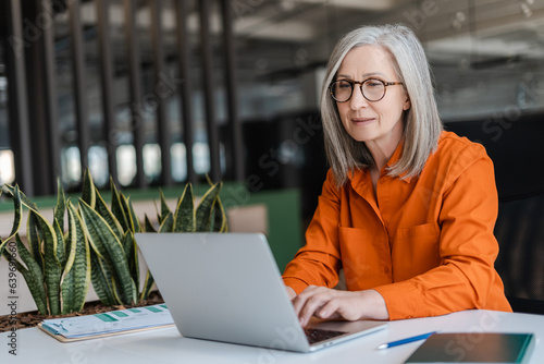 Fotografie, Obraz Confident serious mature businesswoman wearing stylish eyeglasses, orange shirt using laptop working online in modern office