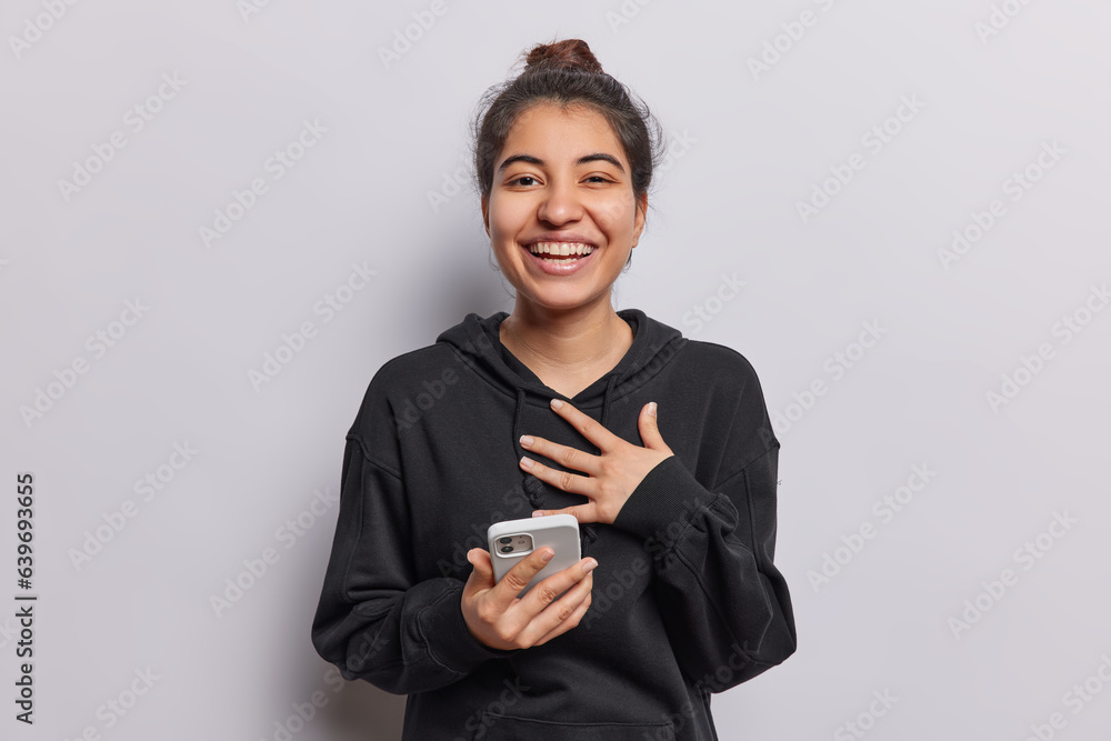Positive sincere young Iranian female model happy to read pleasant news holds modern smartphone keeps hand on chest expresses joy dressed in casual black sweatshirt isolated over white background. Positive sincere young Iranian female model happy to read pleasant news holds modern smartphone keeps hand on chest expresses joy dressed in casual black sweatshirt isolated over white background.