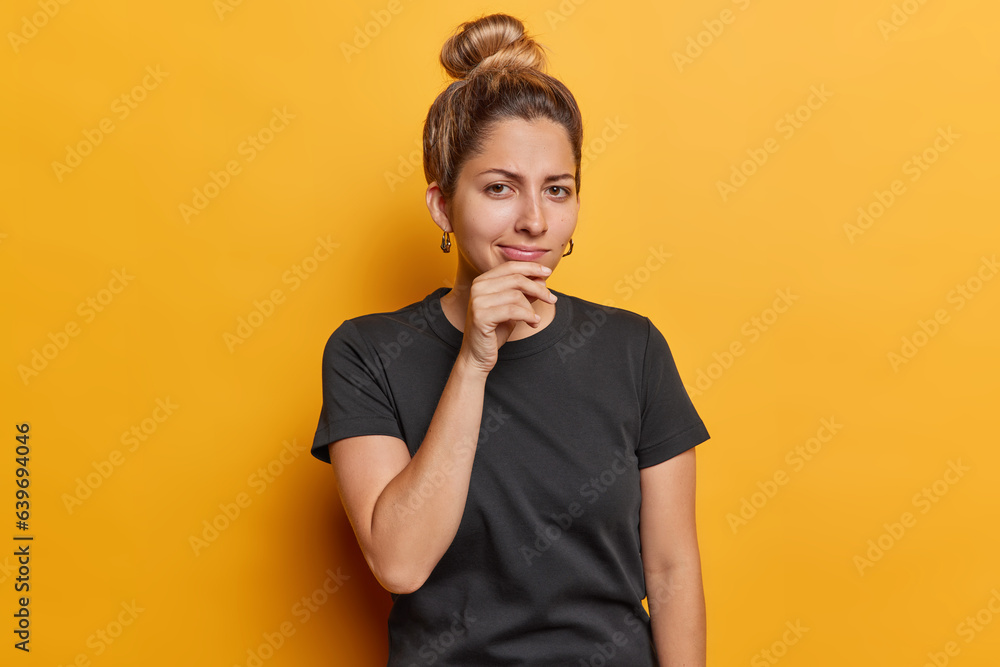 Indoor shot of serious young European woman keeps hand on chin concentrated at camera has attentive gaze wears casual black t shirt isolated over vivid yellow background thinks about something Indoor shot of serious young European woman keeps hand on chin concentrated at camera has attentive gaze wears casual black t shirt isolated over vivid yellow background thinks about something