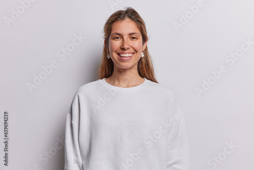 Portrait of cheerful long haired woman smiles toothily being in good mood dressed in casual sweatshirt has piercing in nose isolated over white background. People postive emotions and feelings concept