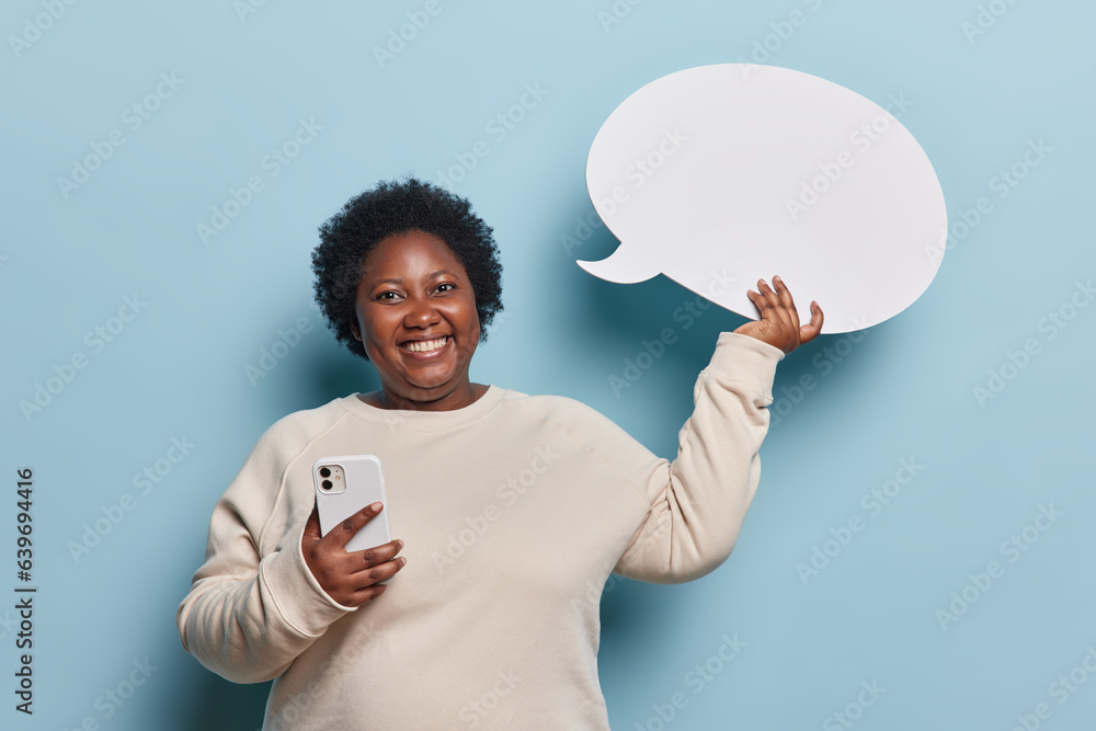 Glad obese dark skinned woman with short curly hair holds smartphone and communication bubble eagerly awaiting for your advertisement wears casual jumper isolated over blue background. Idea concept Glad obese dark skinned woman with short curly hair holds smartphone and communication bubble eagerly awaiting for your advertisement wears casual jumper isolated over blue background. Idea concept