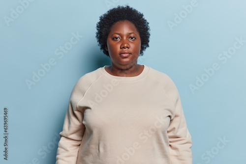 Portrait of serious dark skinned overweight young woman looks directly at camera dressed in sweatshirt concentrated at camera poses against blue background. Human face expressions and emotions