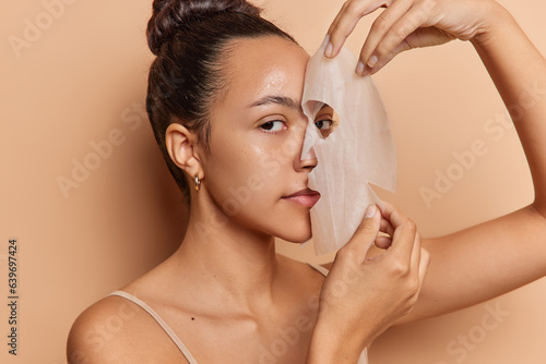 Beauty and cosmetology concept. Studio sideway shot of young pretty cheerful European lady standing isolated in centre on beige background putting off sheet face mask after skin care procedure