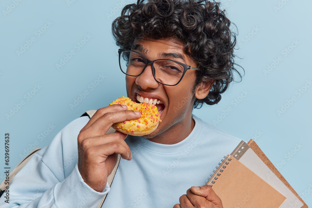 Student lifestyle. Studio photo of young Hindu male wearing casual sweater standing in centre isolated on blue background with bag on shoulder eating delicious doughnut holding books and notebooks Student lifestyle. Studio photo of young Hindu male wearing casual sweater standing in centre isolated on blue background with bag on shoulder eating delicious doughnut holding books and notebooks