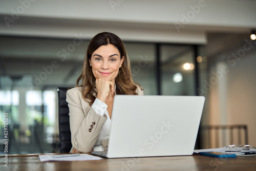 Smiling middle aged 40s professional business woman in office, portrait. Happy mature mid age professional businesswoman executive wearing suit looking at camera at work with laptop sitting at desk. © insta_photos