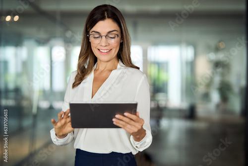 Smiling mid aged mature professional business woman bank manager, 40s female executive, entrepreneur holding fintech tab digital tablet corporate digital technology device standing in office at work.