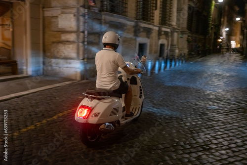 Man rides Italian motor scooter rides down small roman street at night in Rome, Italy 