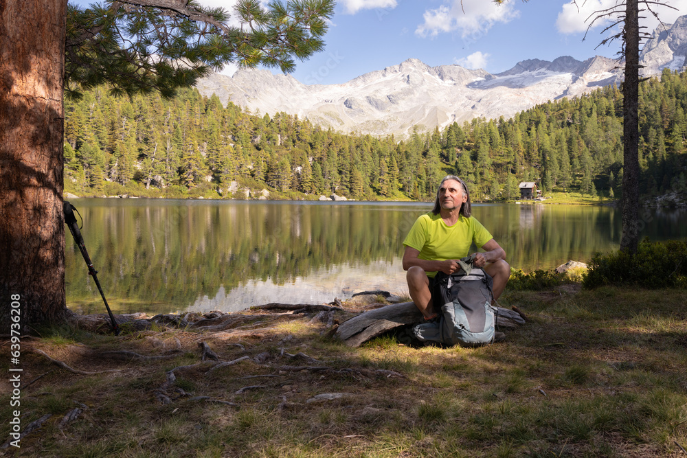Adult gray-haired man with backpack sitting on the shore with his back to a mountain lake with a beautiful reflection Adult gray-haired man with backpack sitting on the shore with his back to a mountain lake with a beautiful reflection