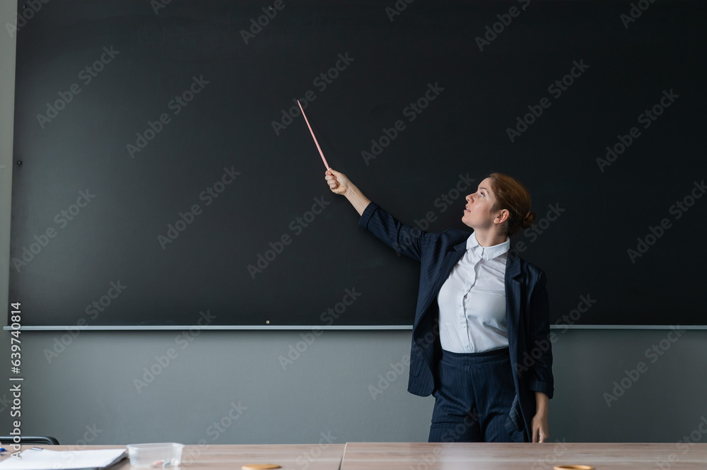Red-haired caucasian woman in a trouser suit. Smiling female teacher with a pointer at the blackboard. Red-haired caucasian woman in a trouser suit. Smiling female teacher with a pointer at the blackboard.