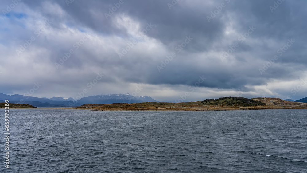 Seascape. A small island with stunted vegetation in the Beagle Strait. In the distance, the Andes mountain range is visible against the cloudy sky. Ripples on the blue water. Argentina. Seascape. A small island with stunted vegetation in the Beagle Strait. In the distance, the Andes mountain range is visible against the cloudy sky. Ripples on the blue water. Argentina.