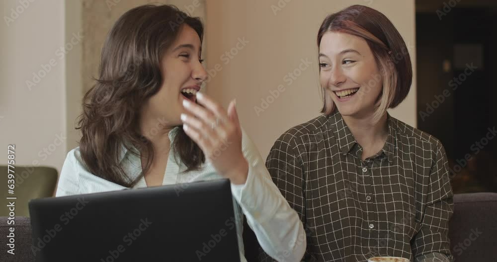 Two female colleagues talking about a project and smiling while resting on the comfy office sofa, camera trucking movement Two female colleagues talking about a project and smiling while resting on the comfy office sofa, camera trucking movement