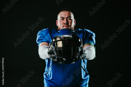 An Asian man with a red beard in a blue american football uniform holds a helmet on a black background.