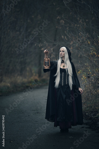 a blonde witch with long hair in a black hoodie stands with an authentic lantern on the road looking into the distance