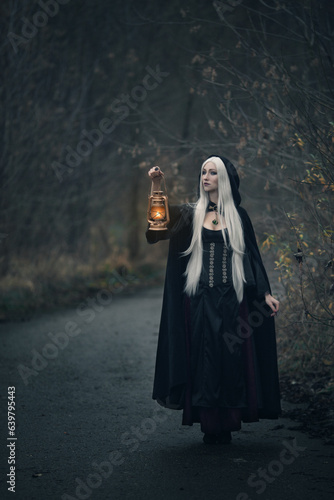 a blonde witch with long hair in a black hoodie stands with an authentic fire lantern on the road looking into the distance