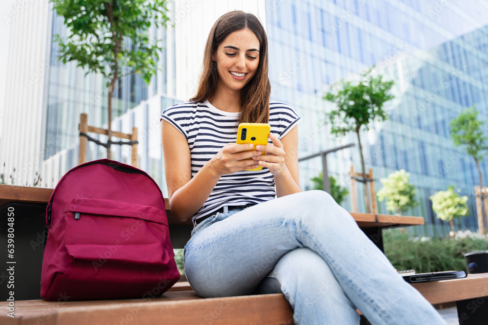 Happy hispanic student girl using yellow cell phone outdoors at University campus. Smiling young woman having fun with online app Happy hispanic student girl using yellow cell phone outdoors at University campus. Smiling young woman having fun with online app
