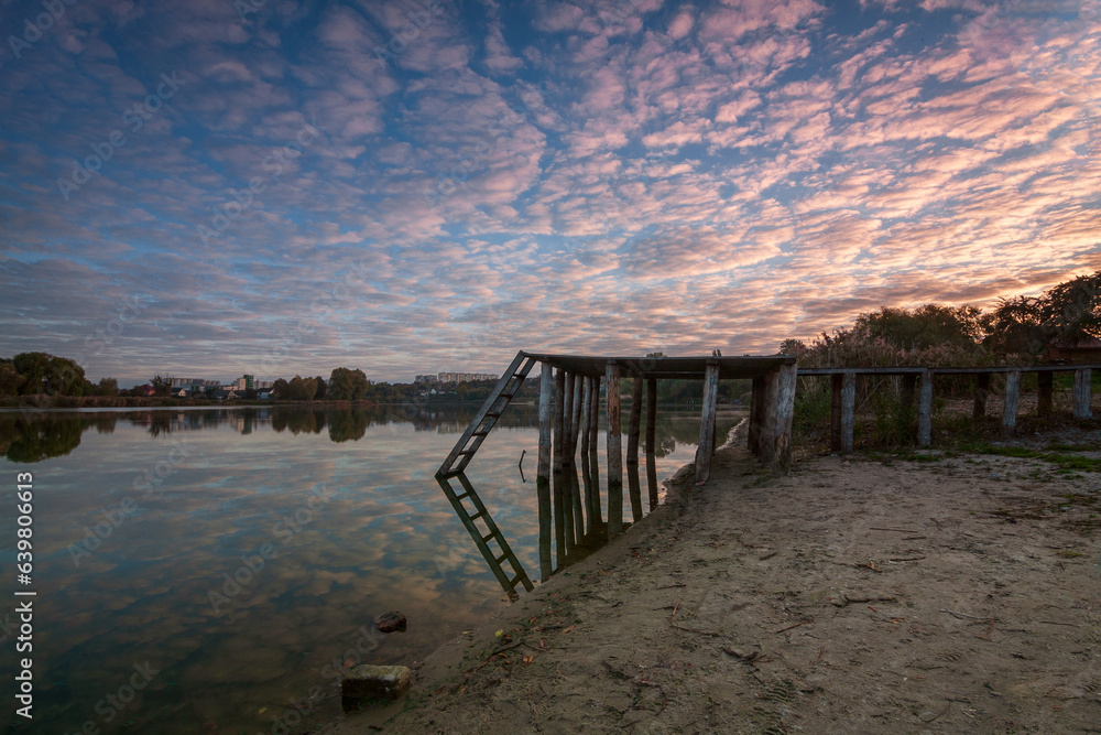 Autumn landscape on the river at dawn, where the sun is rising on the horizon and the sky is pink and blue with white clouds and a bridge, and in the distance reeds and trees