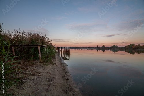 Autumn landscape on the river at dawn, where the sun is rising on the horizon and the sky is pink and blue with white clouds and a bridge, and in the distance reeds and trees