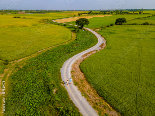 a couple of men and women on vacation in Thailand walking on a curved winding countryside road in the middle of green rice paddy fields in Central Thailand Suphanburi region, drone aerial view