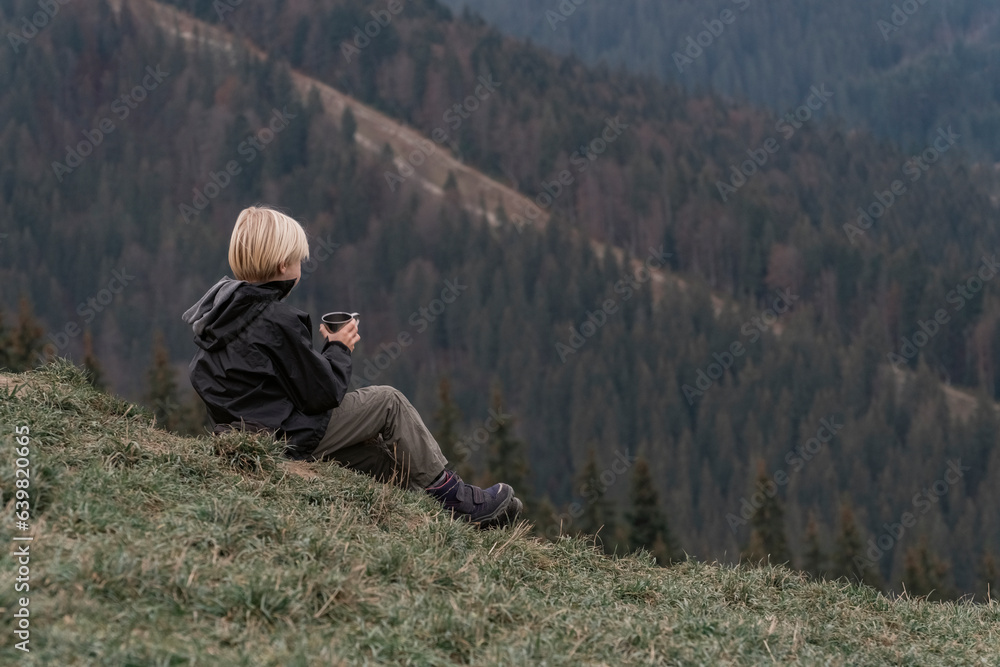 Blond boy in mountains resting on hillside with cup of tea. Hiking with child. Active recreation or vacation Blond boy in mountains resting on hillside with cup of tea. Hiking with child. Active recreation or vacation