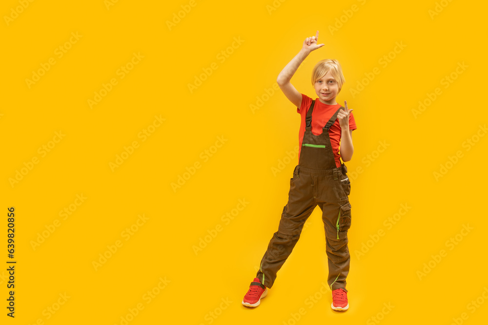 Full-length portrait of boy in overalls pointing with his fingers up. Copy space. Place to advertise Yellow background Full-length portrait of boy in overalls pointing with his fingers up. Copy space. Place to advertise Yellow background