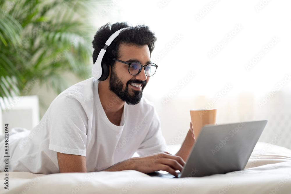 Young Indian Guy Wearing Headphones Resting In Bed With Laptop And Coffee Young Indian Guy Wearing Headphones Resting In Bed With Laptop And Coffee