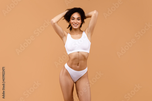 Young fit latin woman in white lingerie posing with hands behind head and smiling at camera on brown background