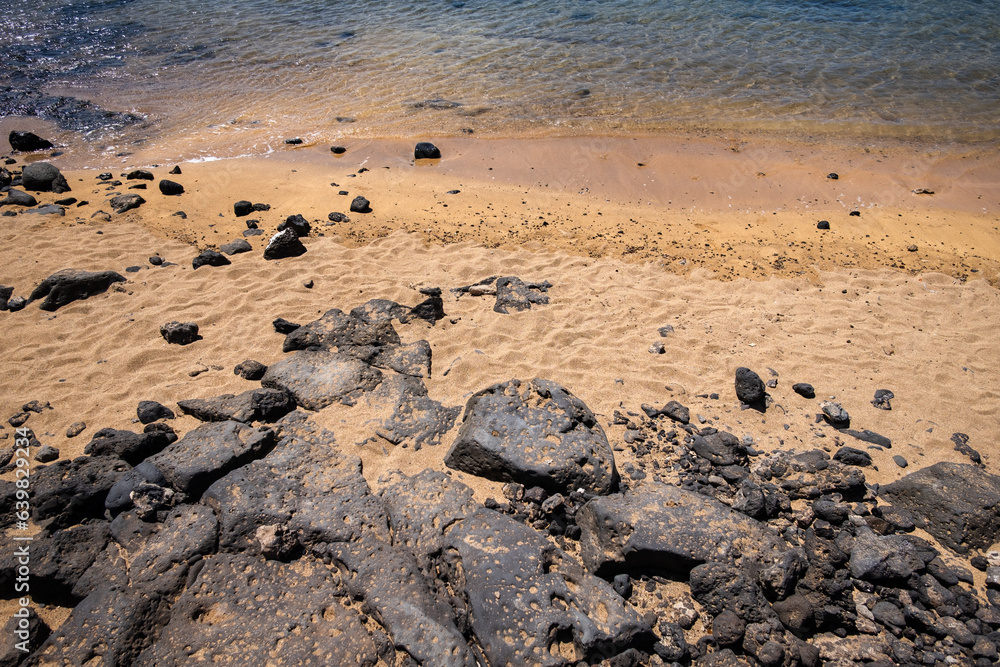 White sand and rocks from the shore. Shore of a white sandy beach with rocks, turquoise sea wets the sand of a beach in Lanzarote. Lanzarote, Canary Islands, Spain White sand and rocks from the shore. Shore of a white sandy beach with rocks, turquoise sea wets the sand of a beach in Lanzarote. Lanzarote, Canary Islands, Spain