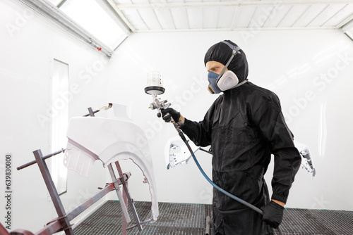 Worker in protective uniform spraying car body part in white paint