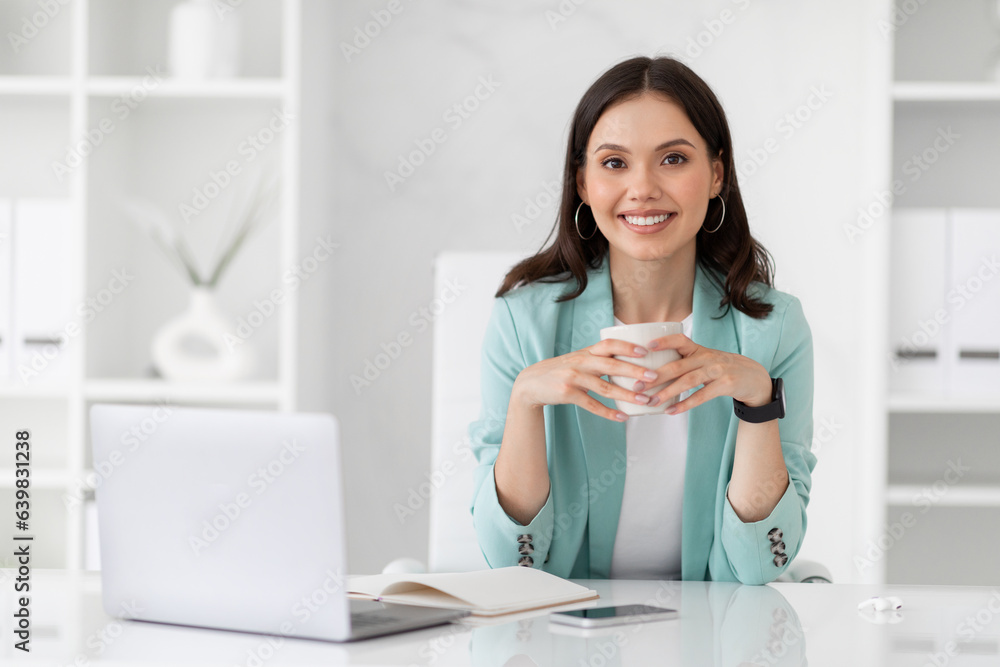 Happy millennial european businesswoman uses computer, drink coffee, enjoy work, relax in white office Happy millennial european businesswoman uses computer, drink coffee, enjoy work, relax in white office