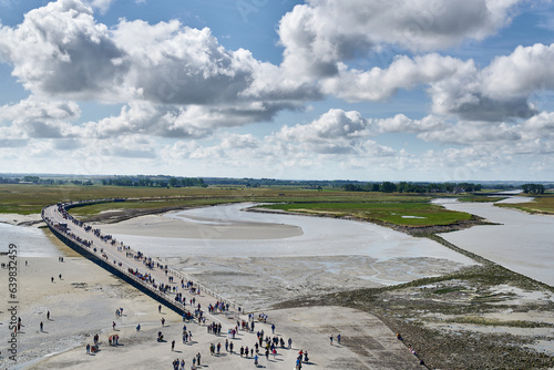 Wooden walkway full of people crossing a bay without water with a green forest in the background, water and the sky covered with white clouds