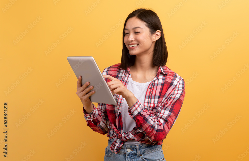 Happy asian lady enjoying her brand new gadget digital tablet, touching pad screen and smiling, yellow background Happy asian lady enjoying her brand new gadget digital tablet, touching pad screen and smiling, yellow background