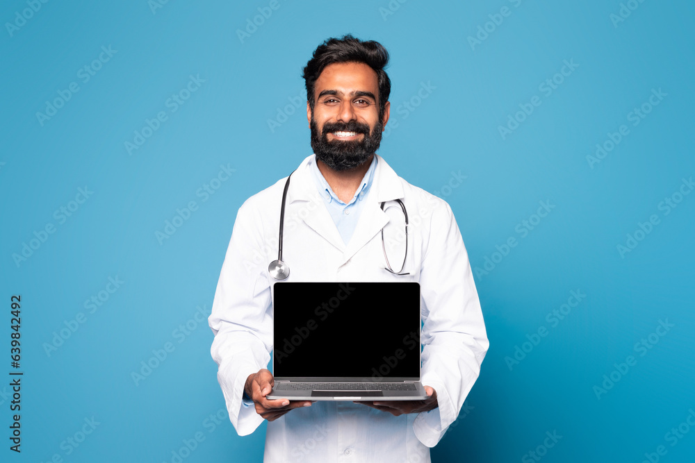 Happy hindu man doctor in white coat with stethoscope showing laptop with blank screen on blue studio background Happy hindu man doctor in white coat with stethoscope showing laptop with blank screen on blue studio background