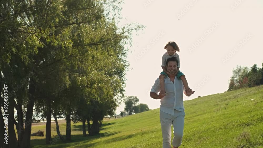 The father carries his son on his shoulders and runs through the grass. The father carries his son on his shoulders and runs through the grass.