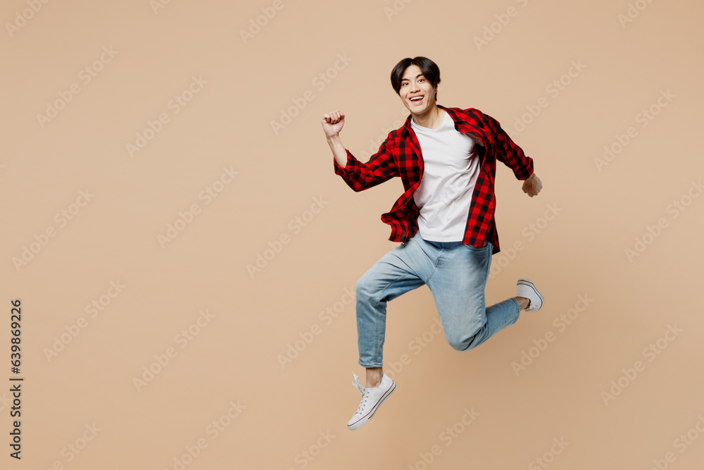 Full body side view young excited cheerful happy man of Asian ethnicity wear red shirt casual clothes jump high run fast look camera isolated on plain pastel light beige background. Lifestyle concept.