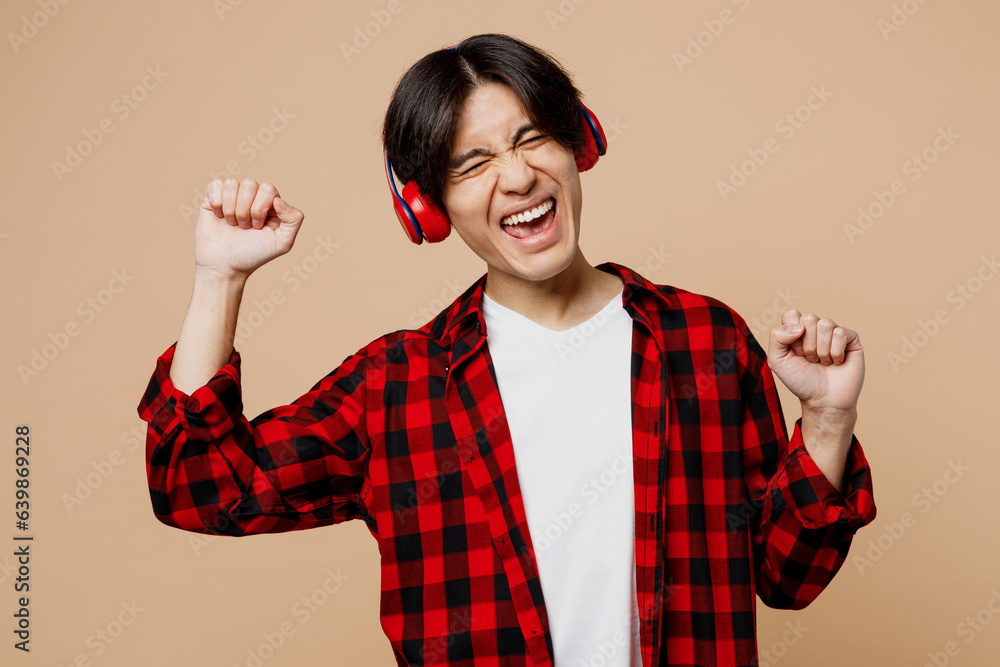 Young excited happy man of Asian ethnicity he wear red shirt casual clothes listen to music in headphones dance raise up hands isolated on plain pastel light beige background studio Lifestyle concept Young excited happy man of Asian ethnicity he wear red shirt casual clothes listen to music in headphones dance raise up hands isolated on plain pastel light beige background studio Lifestyle concept