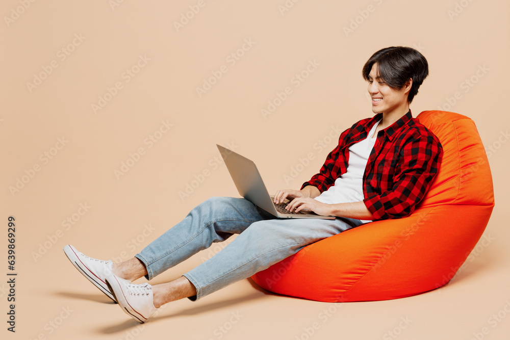 Full body young IT man of Asian ethnicity wear red shirt casual clothes sit in bag chair hold use work on laptop pc computer isolated on plain pastel light beige background studio. Lifestyle concept. Full body young IT man of Asian ethnicity wear red shirt casual clothes sit in bag chair hold use work on laptop pc computer isolated on plain pastel light beige background studio. Lifestyle concept.