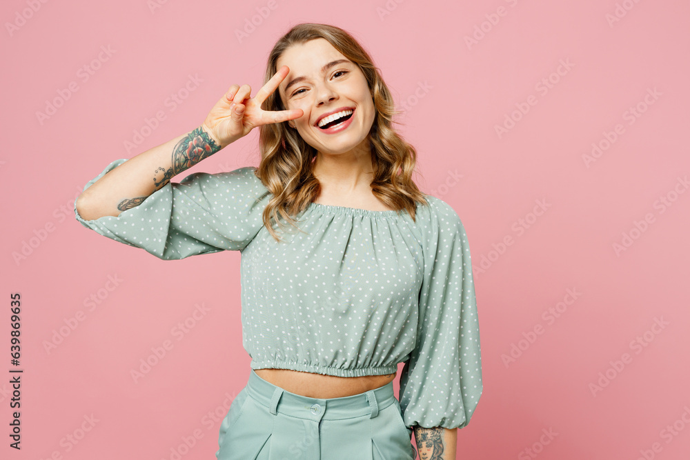 Young smiling cheerful fun happy woman she wear casual clothes look camera show cover eye with victory gesture v-sign isolated on plain pastel light pink background studio portrait. Lifestyle concept. Young smiling cheerful fun happy woman she wear casual clothes look camera show cover eye with victory gesture v-sign isolated on plain pastel light pink background studio portrait. Lifestyle concept.