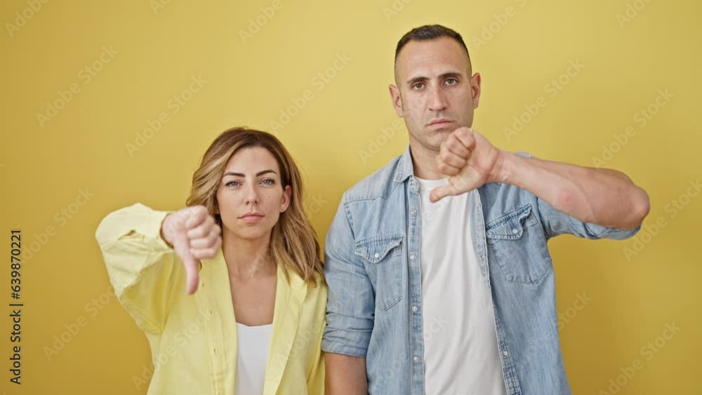 Man and woman couple standing with serious face doing thumb down gesture over isolated yellow background Man and woman couple standing with serious face doing thumb down gesture over isolated yellow background
